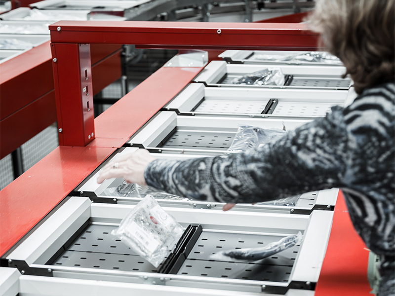 Eine Person legt einen in Kunststoff verpackten Artikel auf ein Split Tray Sorter-Fördersystem mit mehreren Fächern in einer Industrie- oder Lagerumgebung. Die Umgebung scheint automatisiert und organisiert zu sein.
