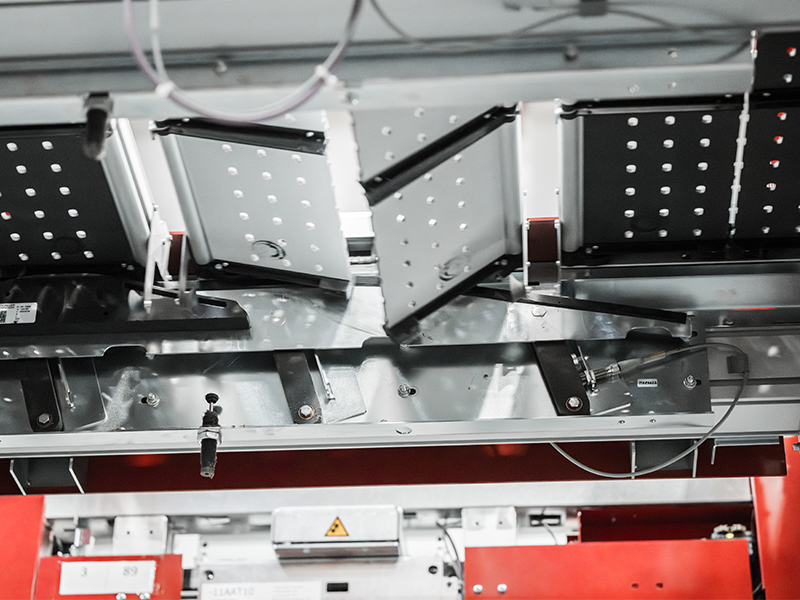 Nahaufnahme einer modernen Industriemaschine, möglicherweise Teil eines Split Tray Sorter-Systems, mit metallischen Komponenten, Verdrahtung, Lochblechen und roten Strukturteilen, die Elemente eines fortschrittlichen automatisierten Fertigungsprozesses hervorheben.