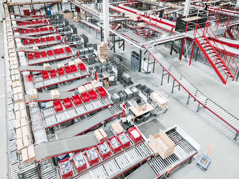 Ein großes Lager mit Förderbändern und einem Split Tray Sorter, der mit Paketen gefüllte rote und graue Behälter transportiert. Kisten sind über die Sortierbänder verstreut, während Metalltreppen und Laufstege über den geschäftigen Maschinen aufragen.