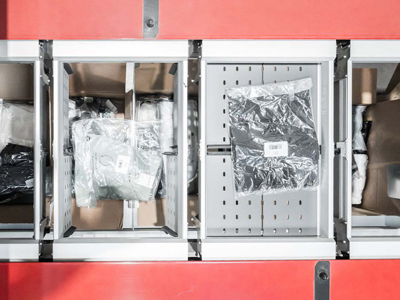 Draufsicht auf drei Fächer in einem roten Split Tray Sorter-Fördersystem, die jeweils durchsichtige Plastikbeutel mit gefalteten Kleidungsstücken und Barcode-Etiketten enthalten.