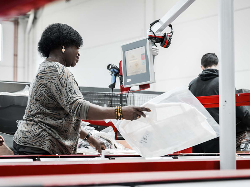 Eine Frau packt Artikel in eine durchsichtige Plastiktüte an einem Arbeitsplatz mit einem Monitor und einem Scanner neben einem Split Tray Sorter in einem scheinbaren Lager oder Vertriebszentrum. Eine weitere Person arbeitet im Hintergrund.
