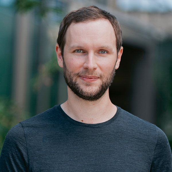 Ein Mann mit kurzen braunen Haaren und Bart, der ein dunkles Hemd trägt, steht im Freien und lächelt in die Kamera und verkörpert das Vertrauen in die Automatisierung.