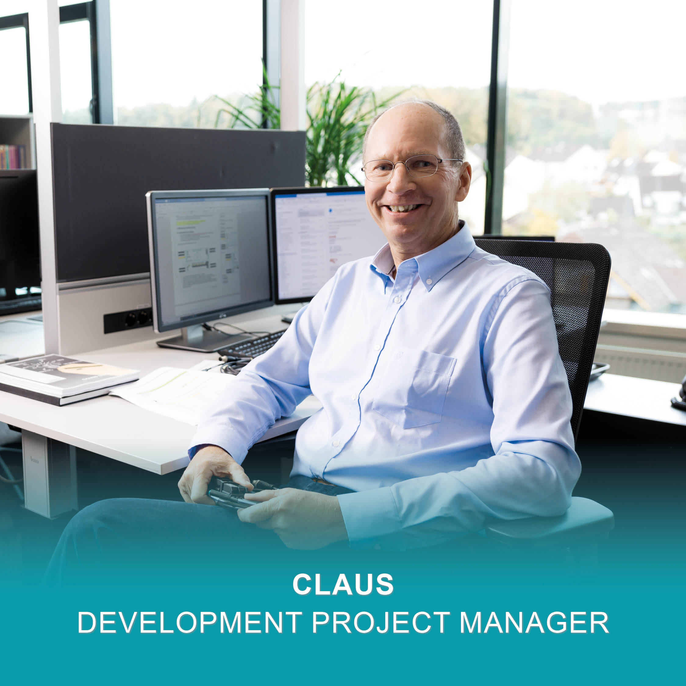 Smiling Development Project Manager in glasses and blue shirt sits at a desk with computer monitors.
