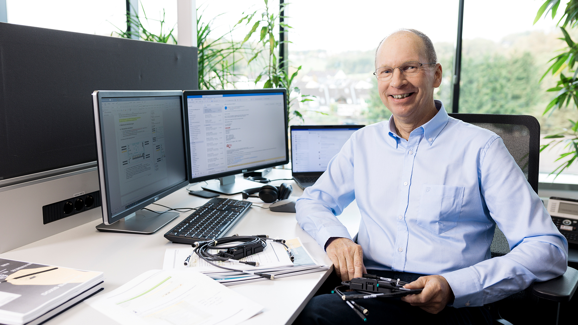 Smiling man sits at a desk with three monitors, a laptop, documents, headphones, and tech equipment in a bright office.