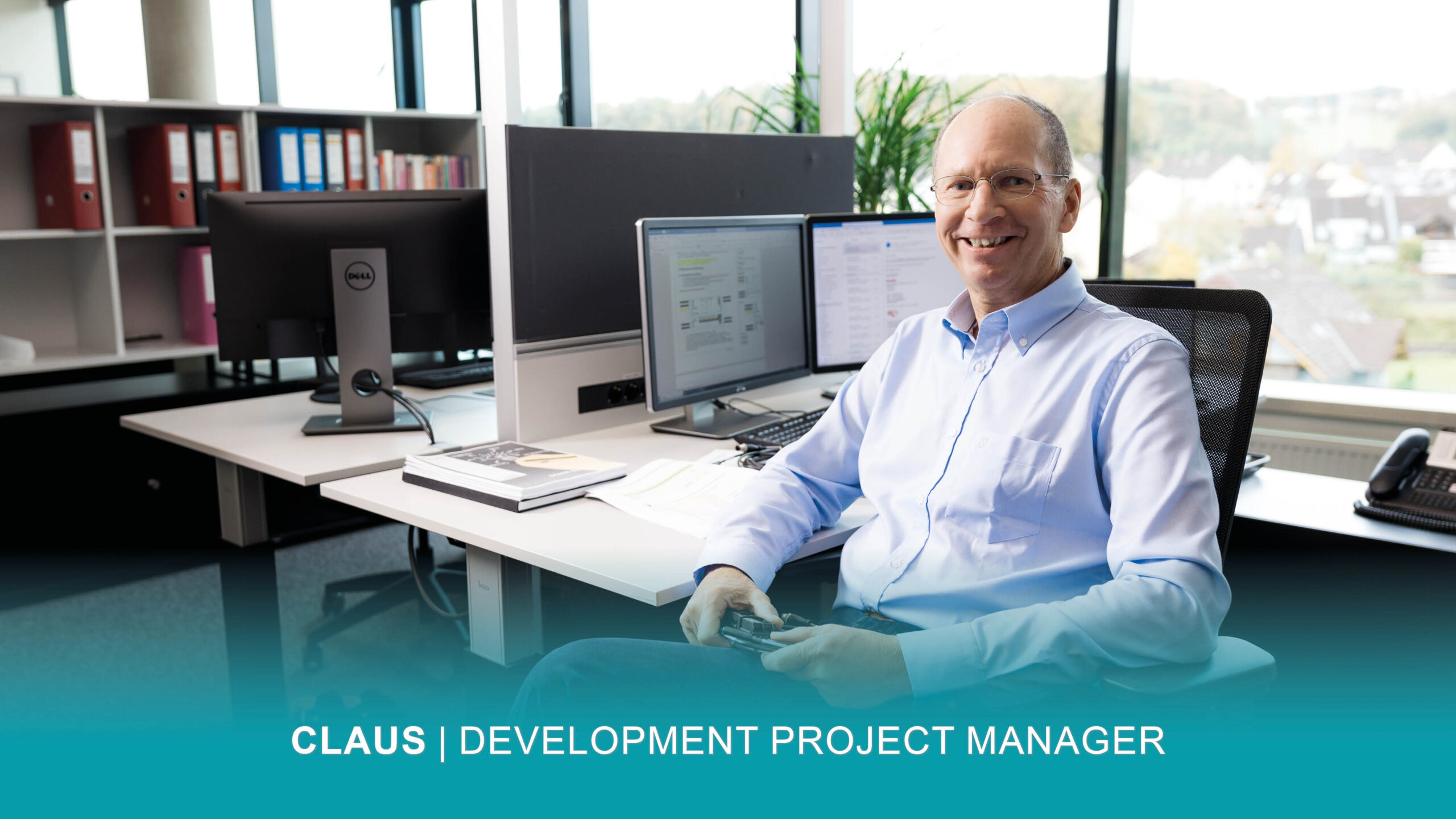 Ein lächelnder Entwicklungsprojektmanager sitzt an einem Schreibtisch mit zwei Monitoren in einem modernen Büro. Der Text lautet: "CLAUS | ENTWICKLUNGSPROJEKTLEITER.