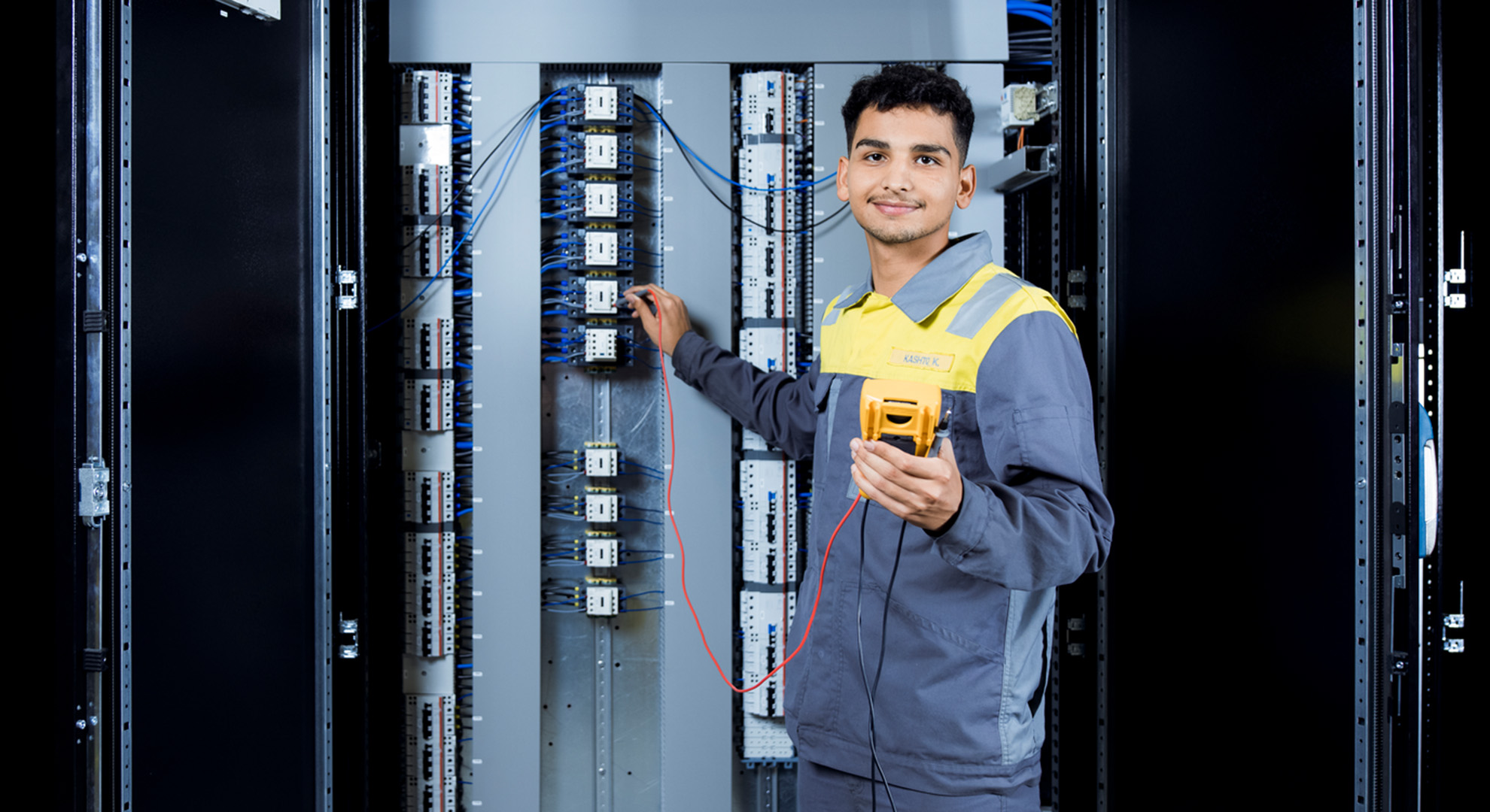 A mechatronic apprentice stands at a switch cabinet and smiles at the camera.