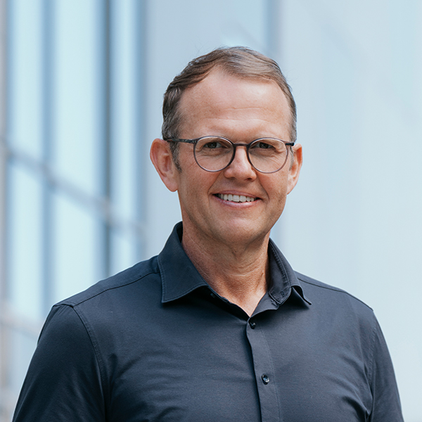 Ein lächelnder Mann mit kurzen braunen Haaren und Brille, der ein dunkles Hemd trägt, steht vor einem verschwommenen Hintergrund mit Glasfenstern.
