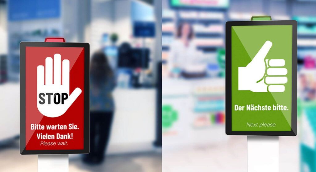 At the pharmacy checkout, one screen displays a red stop sign to wait, while another shows a green thumbs-up to invite the next person forward.