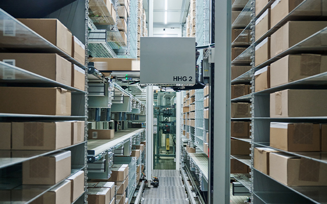 distributioncenter_luxottica_sedico_70 Mit Kartons gefüllte Regalreihen in einem hell erleuchteten, automatisierten Distributionszentrum, in dem sich ein Robotersystem auf einer zentralen Schiene zwischen den Regalen bewegt.