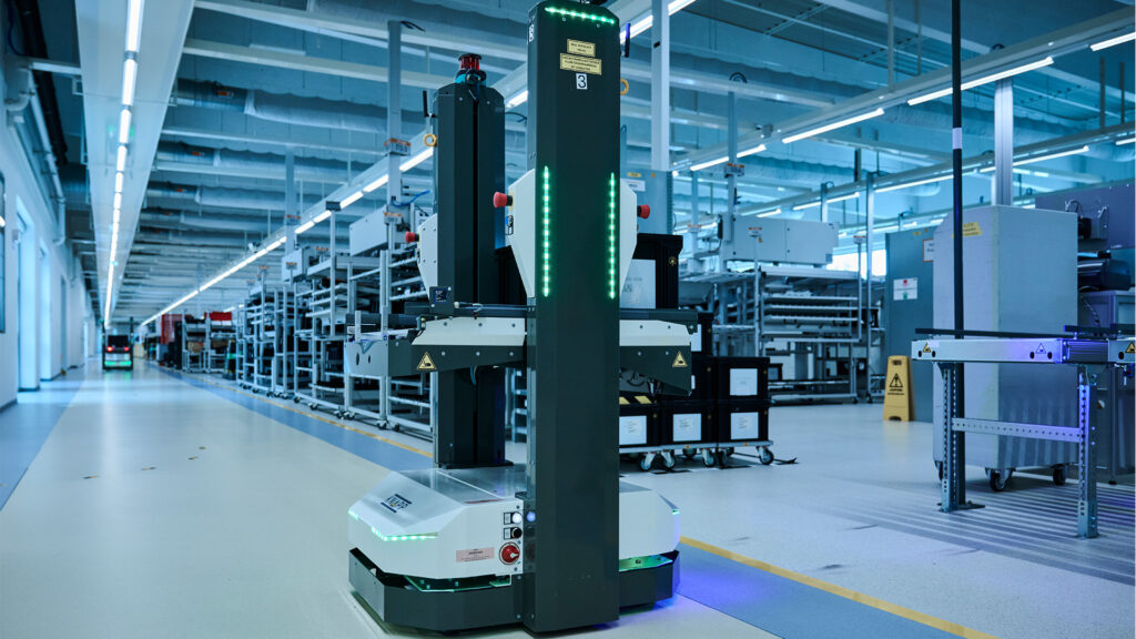 Un AGV AMR con luces LED navega por una fábrica o almacén moderno y bien iluminado, moviéndose entre estanterías y equipos en segundo plano.