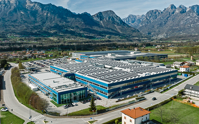 A large industrial building with solar panels on the roof sits at the center of a green valley, surrounded by roads, small houses, fields, and distant mountains under a blue sky.