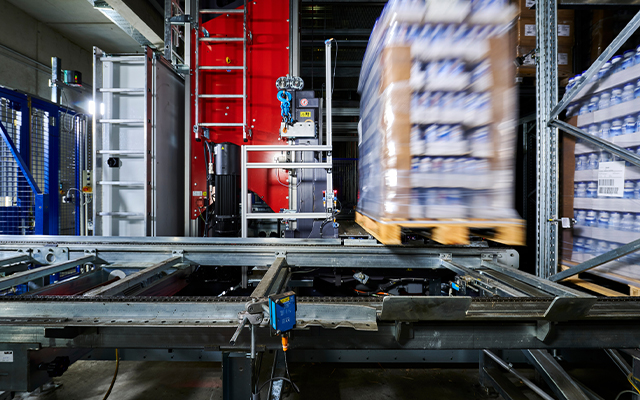 Evo_crane_beitragsbild (RGB): Evo Crane moves a pallet stacked with goods through an automated warehouse, with metal rails and machinery visible in the industrial setting.
