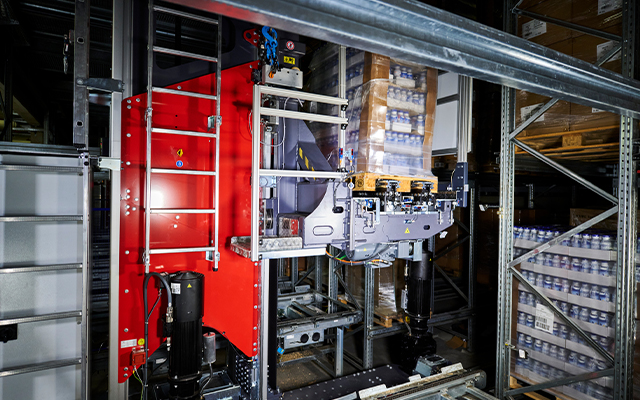 Evo_crane_beitragsbild_2 A robotic (RGB): Evo Crane with a red panel transports stacked boxes on shelves inside a warehouse, showcasing advanced automated logistics and storage systems.