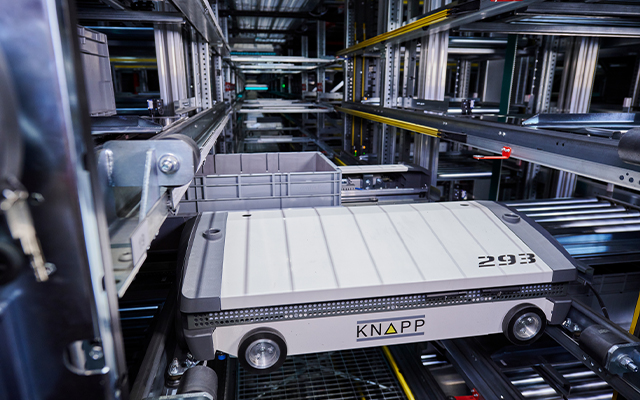 evo_shuttle_arvato_douglas_640x400 Une navette robotisée marquée "KNAPP" transporte un bac de stockage gris à travers un système d'entrepôt automatisé composé de rails et d'étagères métalliques.