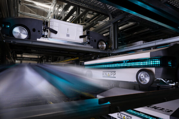 evo_shuttle_technology Des navettes robotisées se déplacent rapidement sur des rails dans un entrepôt de haute technologie, transportant des conteneurs blancs pour les stocker ou les récupérer. Des lumières bleues éclairent leurs trajectoires, mettant en évidence le système d'automatisation avancé.