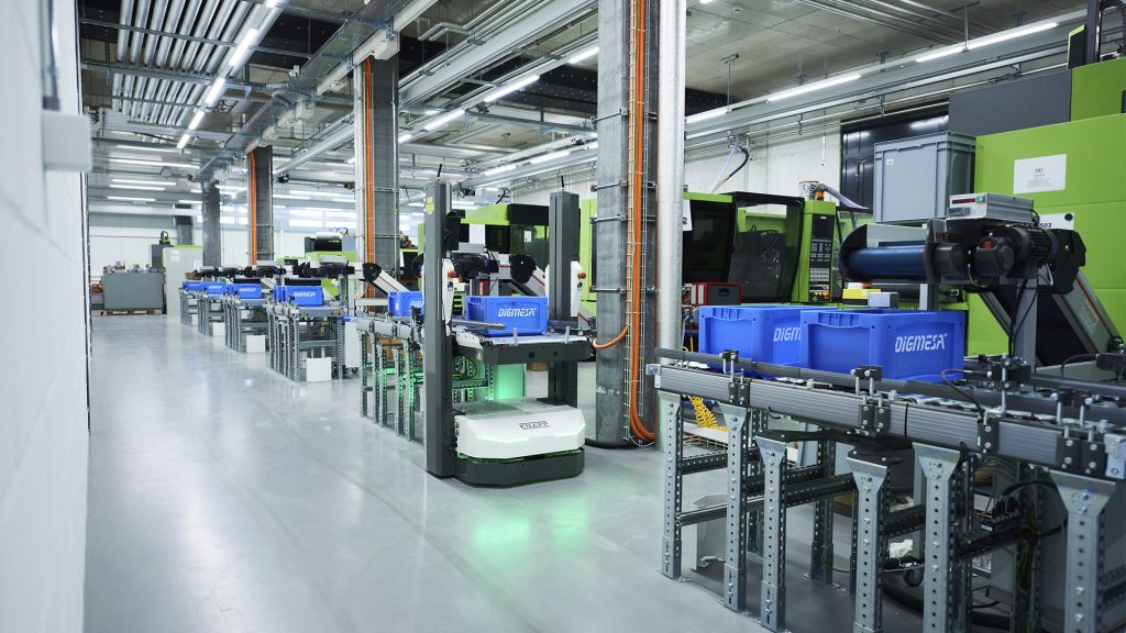 Sistema de transporte en la industria del plástico Frente a las máquinas de moldeo por inyección se encuentra el sistema de transporte con cajas de Digmesa azules. Un RMA entrega una caja.