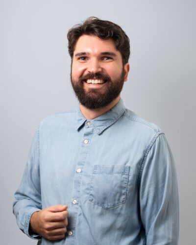 Foto-Armin Un homme souriant, aux cheveux foncés et à la barbe, vêtu d'une chemise bleu clair boutonnée, debout devant un fond uni gris clair.