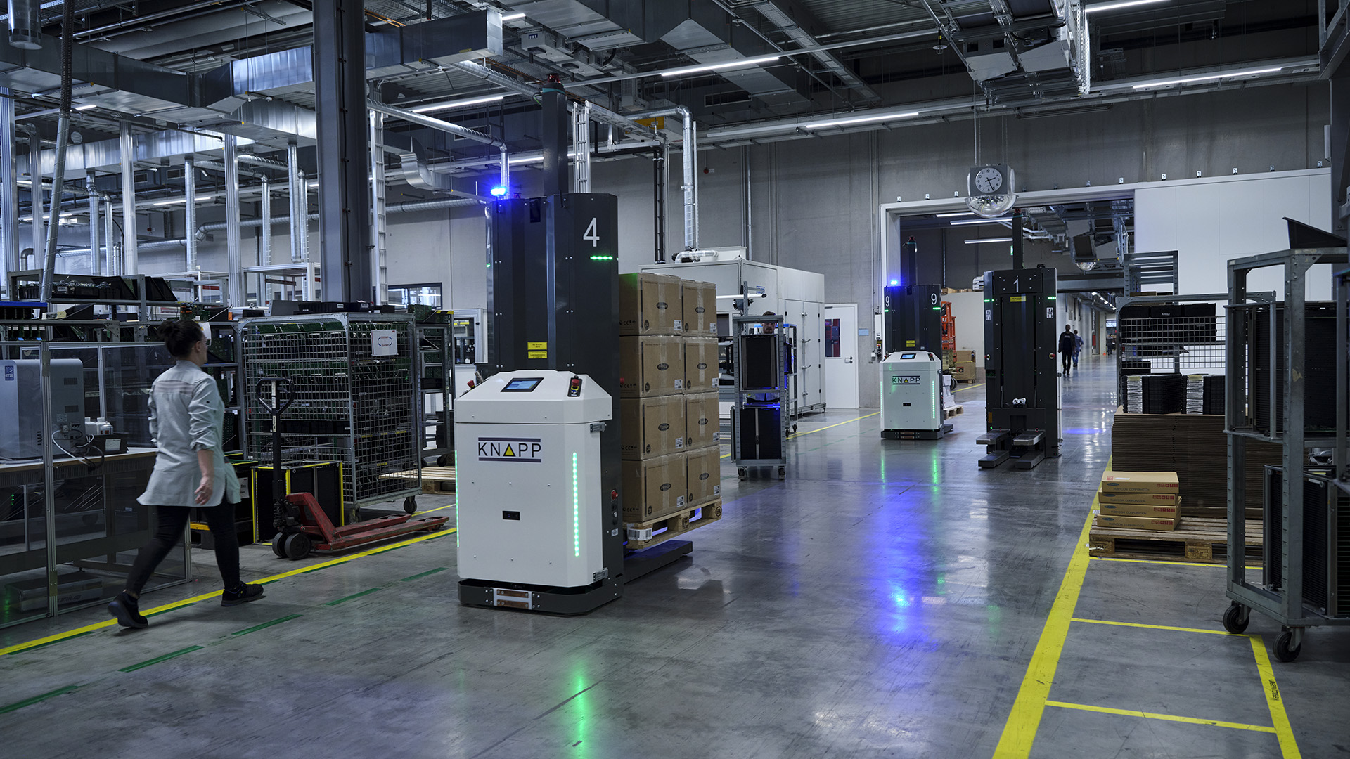 Drei autonome mobile Roboter fahren in einer Lagerhalle, einer transportiert Kartons auf einer Palette.