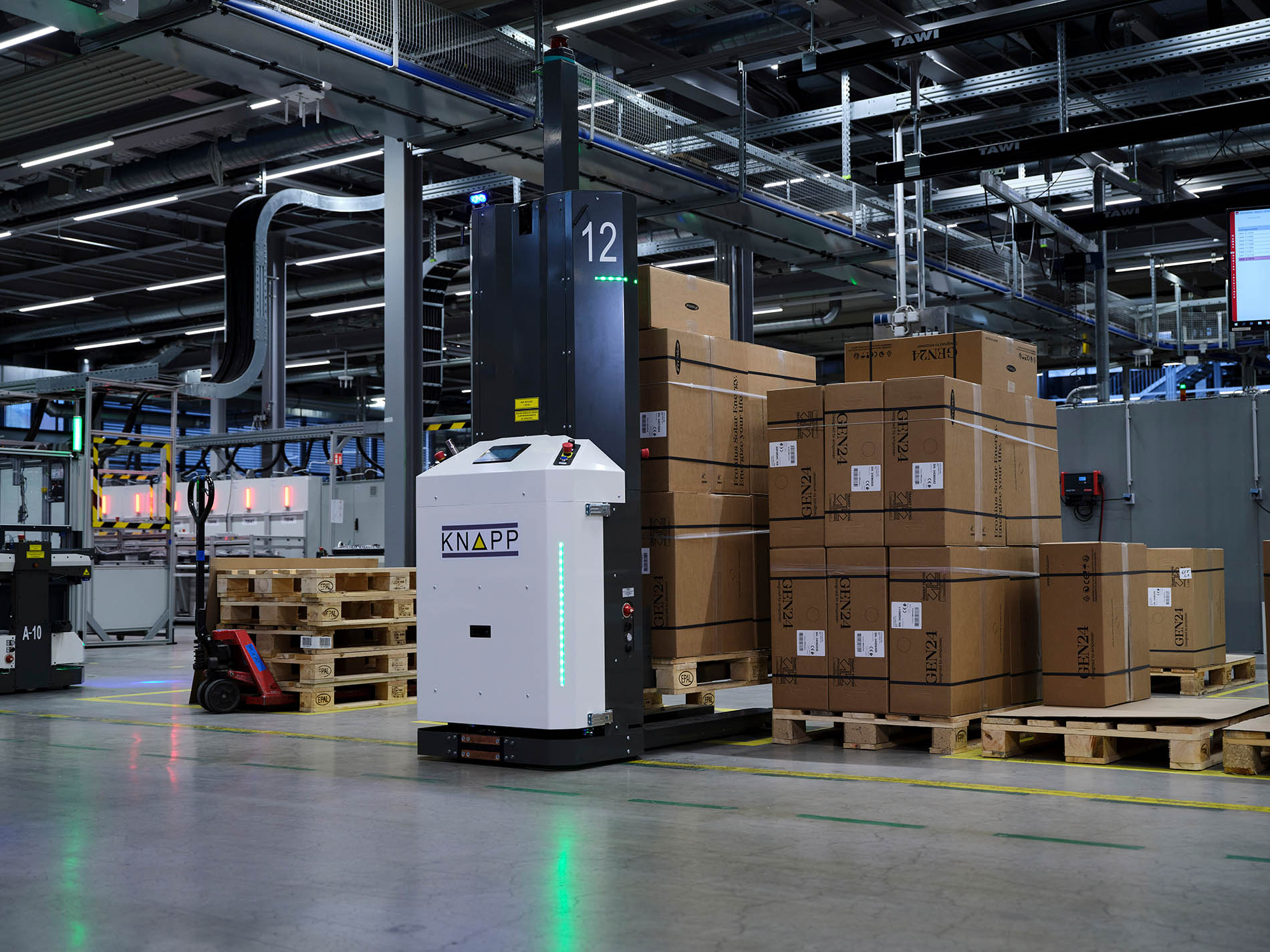 Ein Lagerhaus mit gestapelten Kartons auf Paletten, einem autonomen KNAPP-Roboter und Industrieanlagen unter hellen Deckenleuchten.