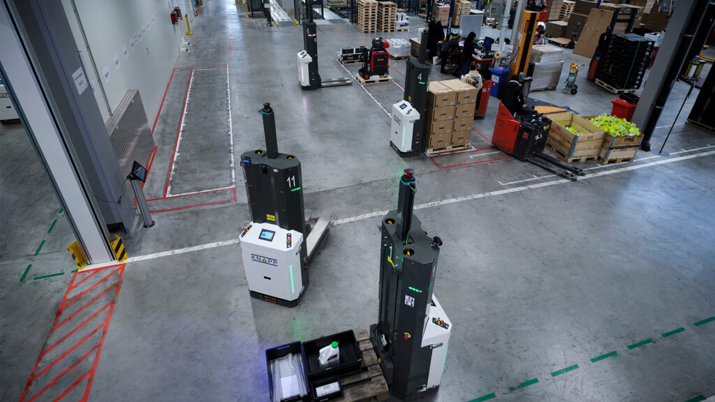 Automatisierte Roboter transportieren Paletten in einem geräumigen Lager mit Arbeitern und gestapelten Kisten im Hintergrund.