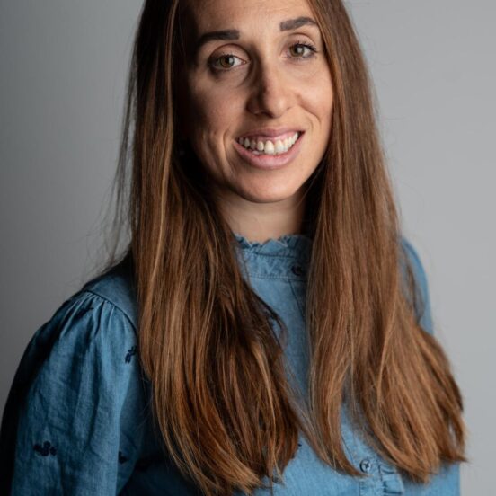Une femme aux longs cheveux bruns, vêtue d'une chemise bleue, sourit à l'appareil photo sur un fond gris uni.