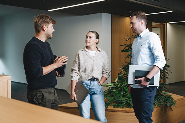 Drei Personen diskutieren über Integrationspartnerschaft in einem modernen Büro, jeder mit einem Laptop oder Tablet, umgeben von Pflanzen und stilvoller Einrichtung.