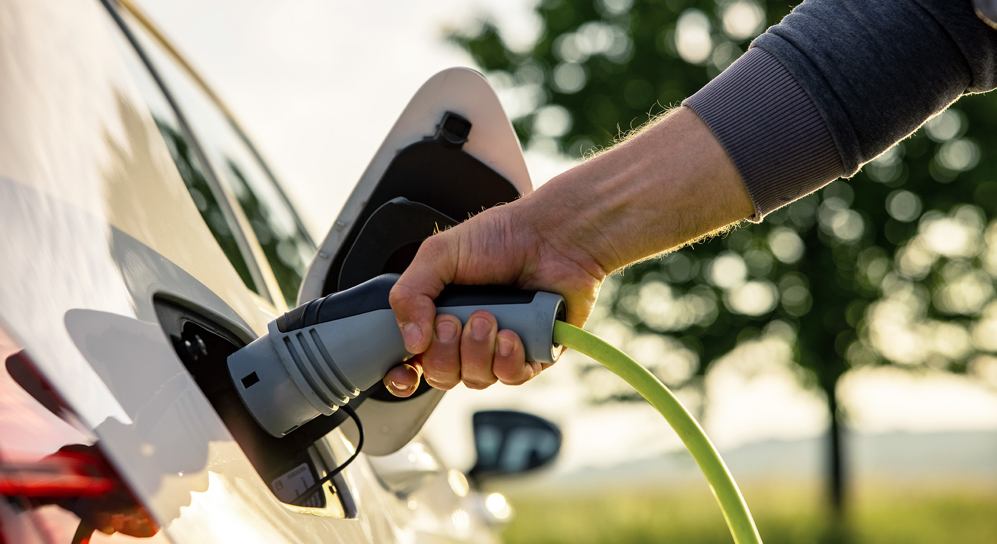 A man’s hand plugs in an electric car for recharging an environmentally friendly car in a green landscape