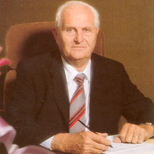 An older man wearing a suit and a striped tie is sitting at a desk and writing on a piece of paper.