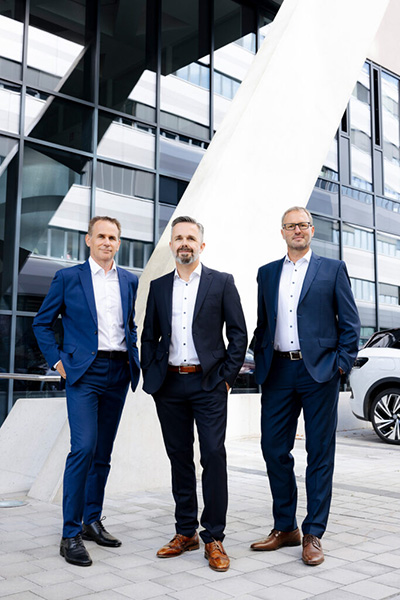 Drei Personen in Business Outfits stehen vor einem modernen Bürogebäude und blicken in unterschiedliche Richtungen.