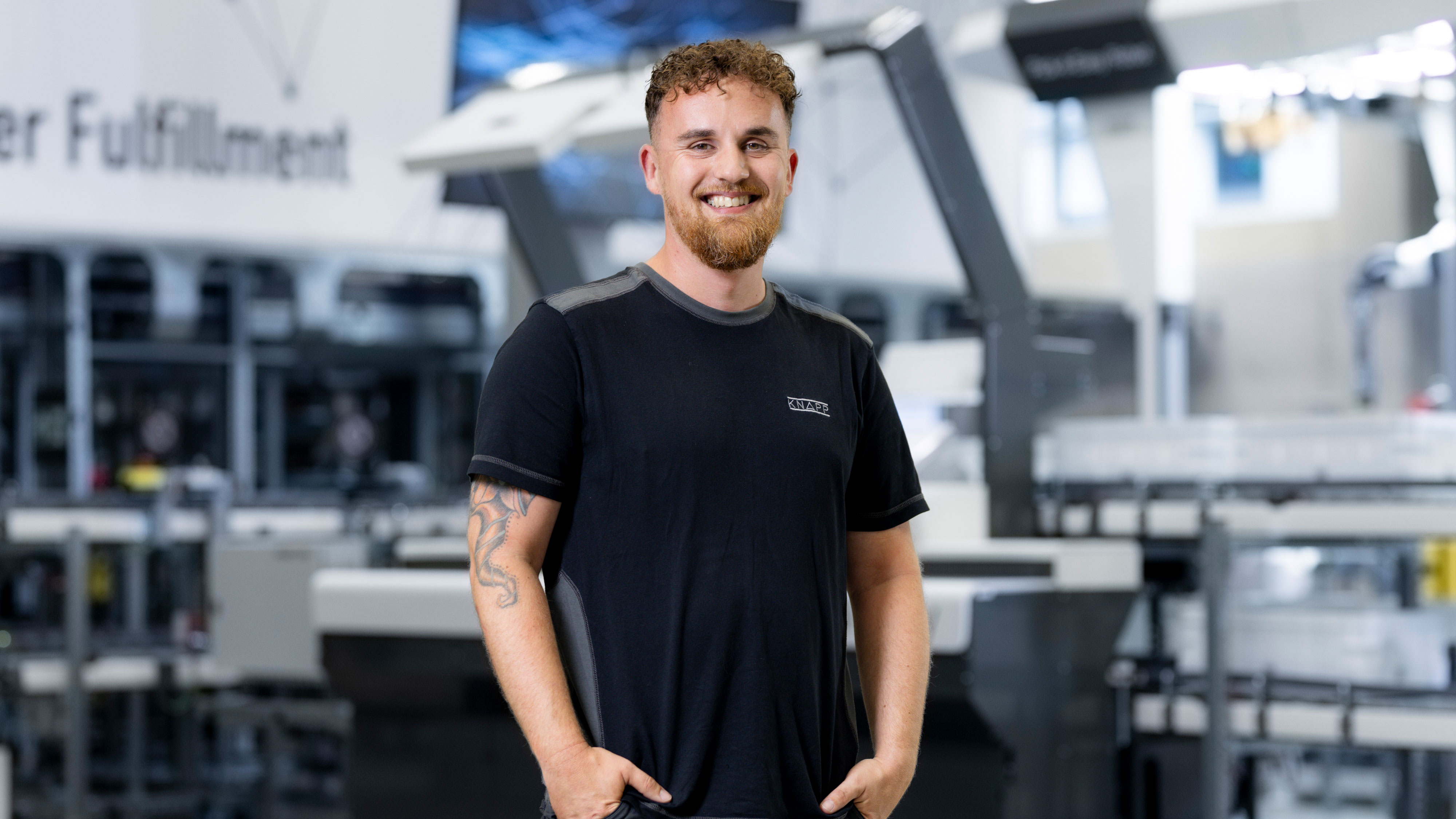 Ein lächelnder Mann mit lockigem Haar und Bart, der ein schwarzes T-Shirt trägt, steht in einer modernen Lagerhalle oder einem Fulfillment-Center inmitten von Industrieanlagen und ist wahrscheinlich mit der Inbetriebnahme von Steuerungen beschäftigt.