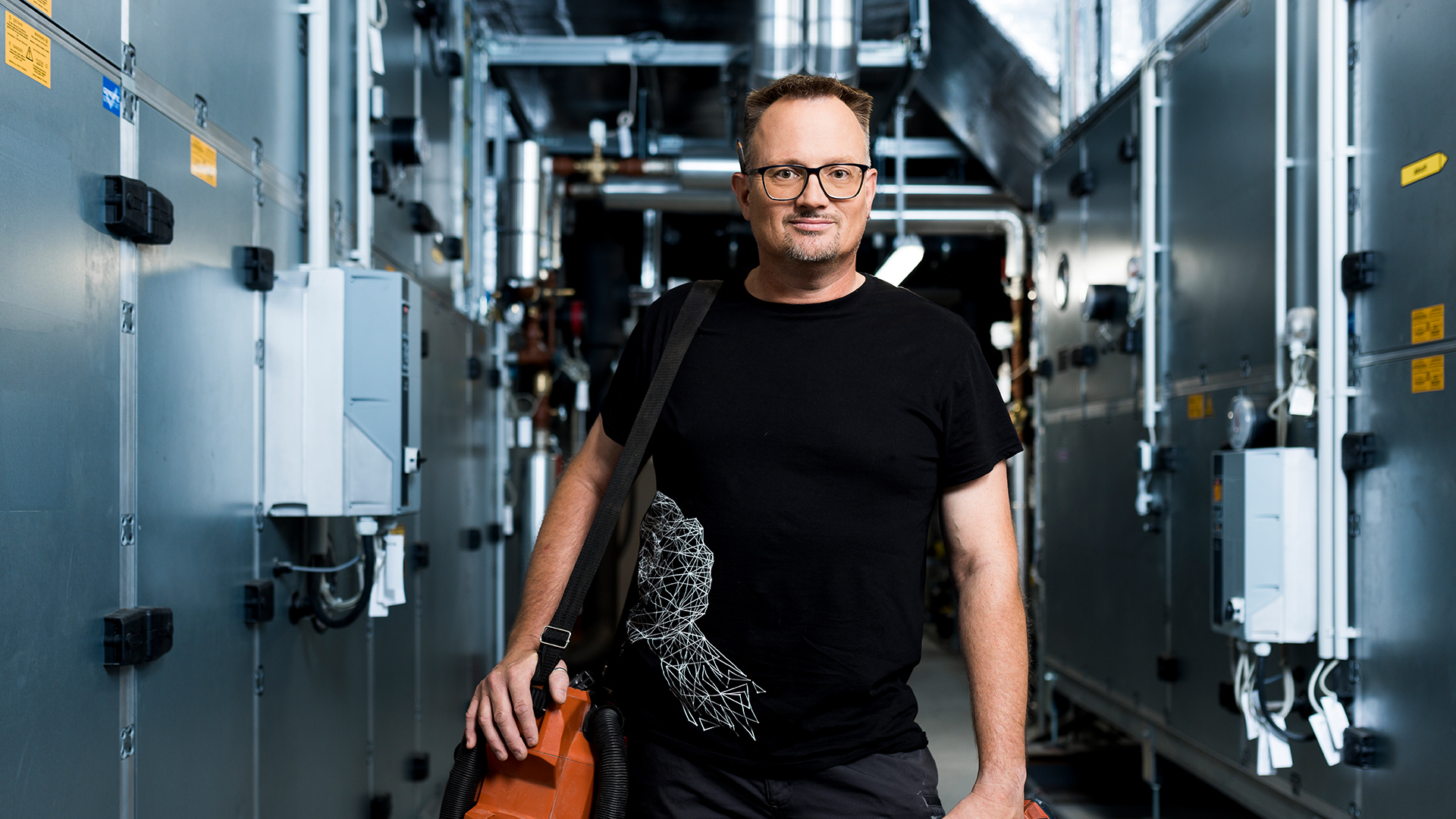 Un homme avec des lunettes et un t-shirt noir se tient dans un couloir industriel, tenant une mallette orange et un sac, entouré de grosses machines.