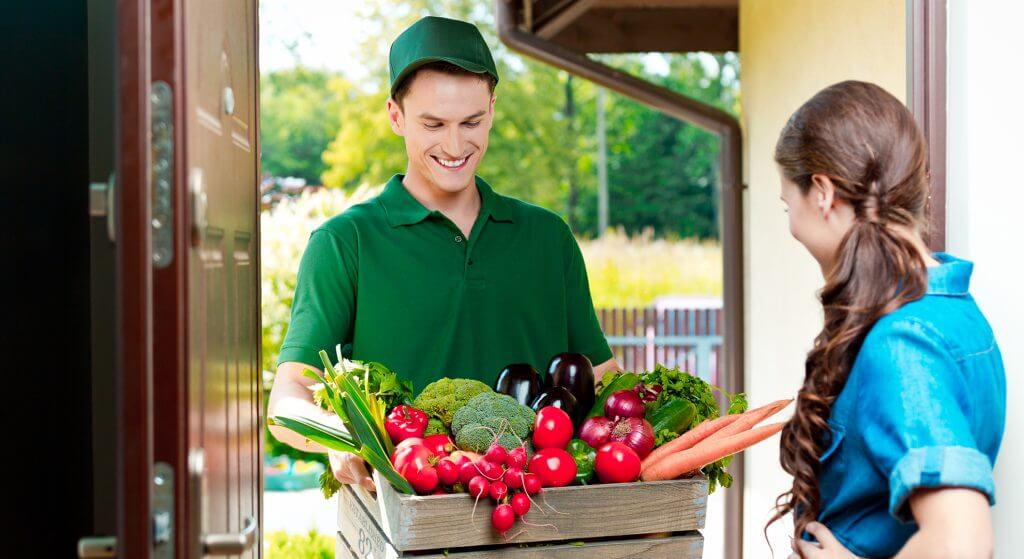 The picture shows a home delivery of groceries. A young delivery man hands a young woman a crate full of fresh vegetables. The woman ordered the groceries online and has them conveniently delivered to her home. The wooden crate holds fresh tomatoes, radishes, onions, eggplants, lettuce, broccoli, zucchini and peppers. The two are looking at the vegetables and smiling.