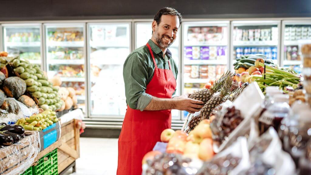 The image shows a male supermarket employee filling shelves with fresh fruit. He is looking directly into the camera and is smiling. At his side are crates with fresh fruits and vegetables, for example, pineapples, apples, peppers, eggplants, cucumbers, zucchinis and so on.