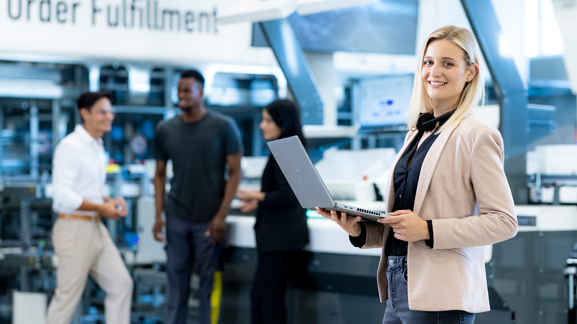 A woman stands with her laptop in hand in front of an installation and smiles at the camera. Three colleagues are seen in the background.