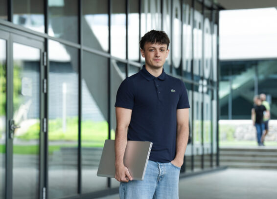 Un jeune homme vêtu d'un polo foncé et d'un jean se tient debout, un ordinateur portable à la main, devant un immeuble aux façades vitrées.