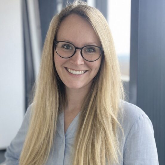 Eine lächelnde Frau mit langen blonden Haaren und Brille, die ein hellblaues Hemd trägt, steht in einem Innenraum an großen Fenstern.