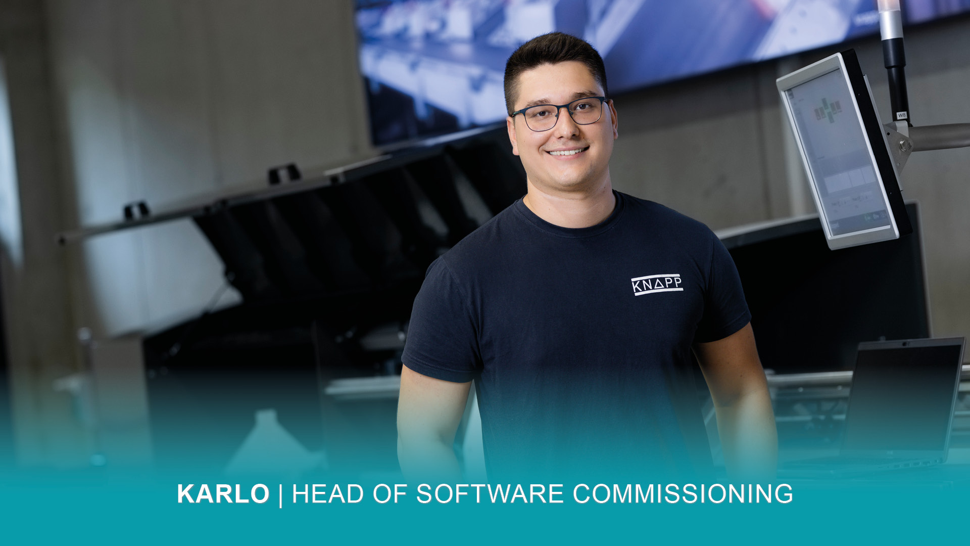 Un hombre sonriente con gafas y camiseta oscura KNAPP en un entorno industrial moderno con pantallas de ordenador. El texto de la parte inferior dice: "KARLO | JEFE DE PUESTA EN SERVICIO DE SOFTWARE".