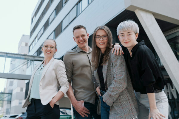 Karriere_Software_Hero-Bild_1920_1080 Vier Personen stehen lächelnd im Freien vor einem modernen Bürogebäude und posieren selbstbewusst - perfekt für ein Fashion Logistik-Teamfoto.