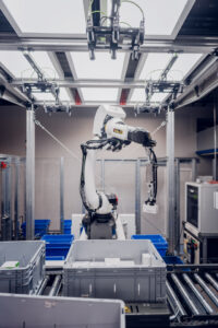 Ein Roboterarm sortiert Gegenstände in graue Behälter – und präsentiert damit Automatisierungslösungen für die gesamte Wertschöpfungskette in einer Industrieautomationsanlage.