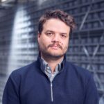Ein Mann mit kurzen braunen Haaren und einem Bart, der einen marineblauen Pullover und ein gestreiftes Hemd trägt, steht in einer Brødrene Dahl-ähnlichen Lagerhalle.