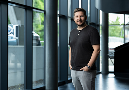 KNAPP CS_2026_076(C)KANIZAJ photography_Christoph Man in a black shirt and gray pants stands smiling in a modern building with large windows and natural light.