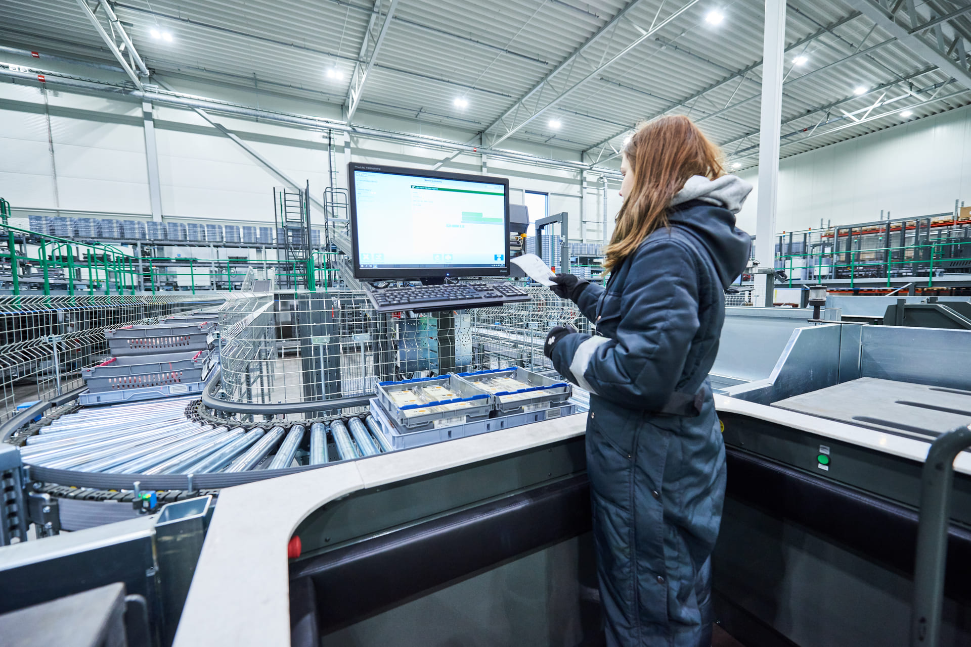 Ein Arbeiter in einem modernen Industriegebäude steht im Overall am Arbeitsplatz, hält ein Papier und betrachtet einen großen Monitor mit Diagrammen - effiziente Automatisierung für die Filialbelieferung im Hintergrund mit Förderbändern und Maschinen.