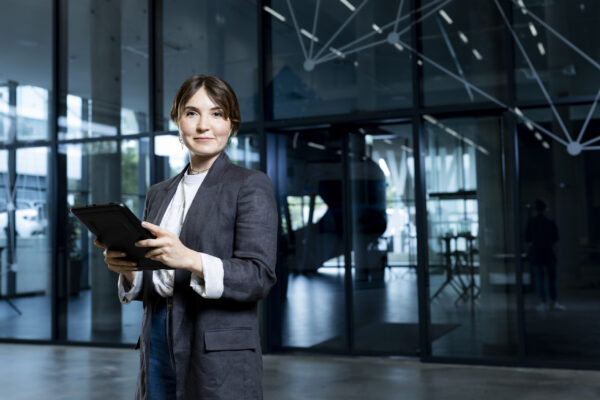 KNAPP-Digital Services 2 (1) Une femme en tenue de travail tenant une tablette se tient avec assurance dans un immeuble de bureaux moderne aux murs de verre et aux motifs géométriques.