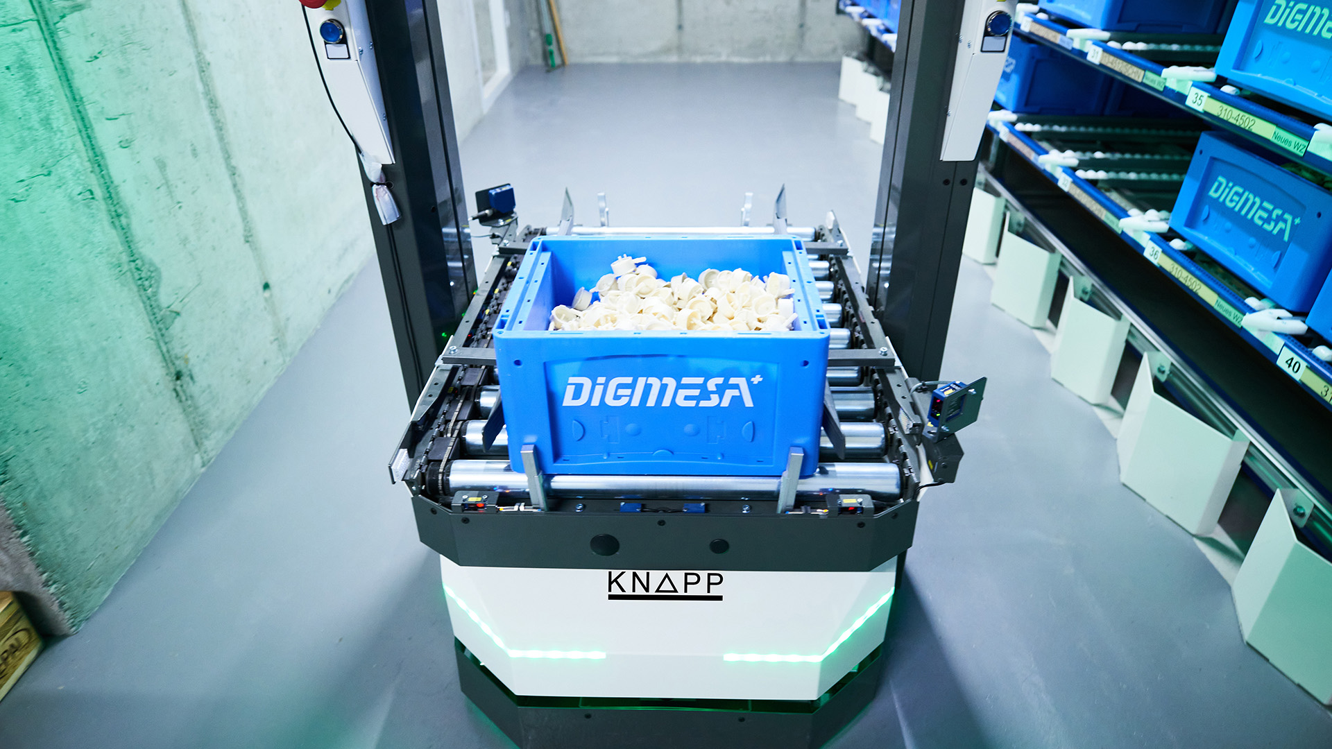Ein autonomer mobiler Roboter fährt mit einer blauen Kiste voller kleiner Kunststoffteile an Regalen vorbei.