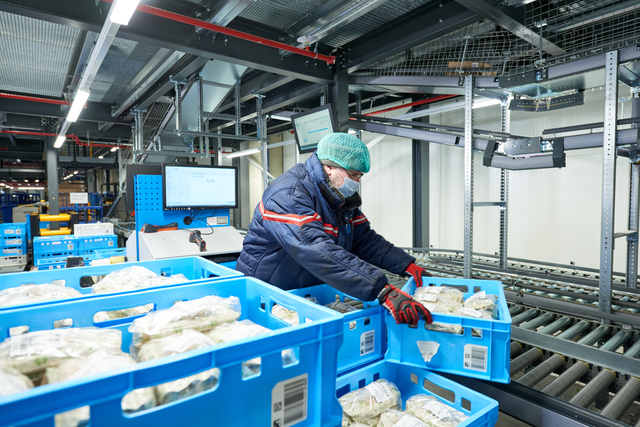 Ein Arbeiter in Winterkleidung bewegt blaue Kisten mit verpackten Lebensmitteln und sorgt für maximale Frische durch maßgeschneiderte Automatisierung im Lager.