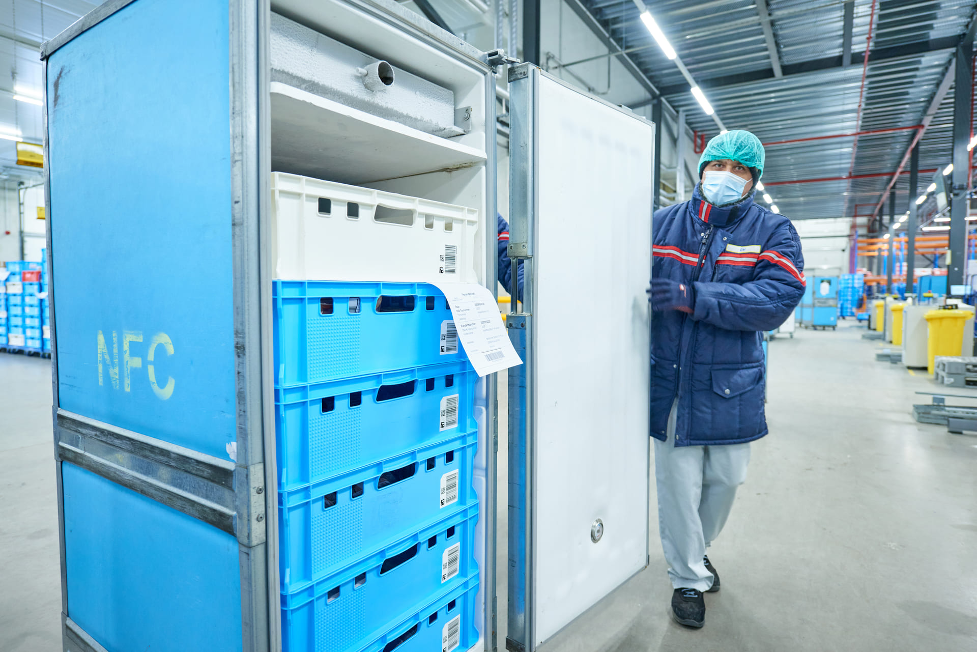 Ein Arbeiter in einer Fabrik, der Maske, Handschuhe und Haarnetz trägt, öffnet einen großen metallenen Lagerbehälter, der mit gestapelten blauen Kisten gefüllt ist - eine Umgebung, die maximale Frische durch maßgeschneiderte Automatisierung gewährleistet.