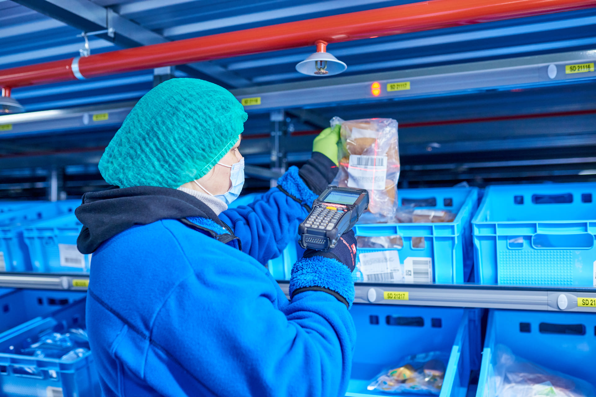 Un ouvrier portant des vêtements de protection bleus, une charlotte verte et un masque facial scanne un article emballé à l'aide d'un lecteur de codes-barres portatif dans un entrepôt doté de bacs de stockage bleus.