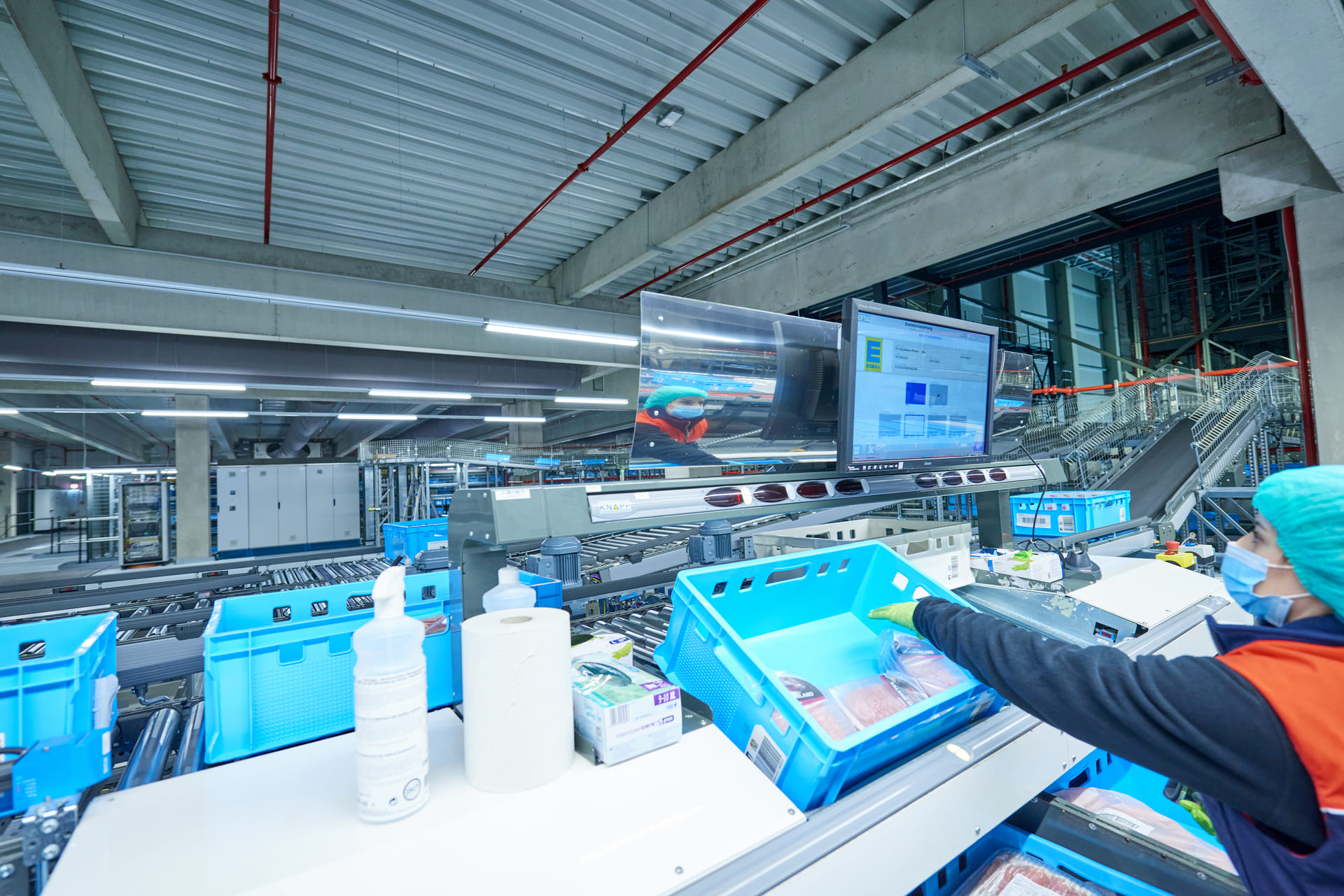 Un travailleur en tenue de protection trie des produits dans des bacs bleus dans un entrepôt automatisé moderne où l'on peut voir des tapis roulants, des moniteurs et des produits de nettoyage.