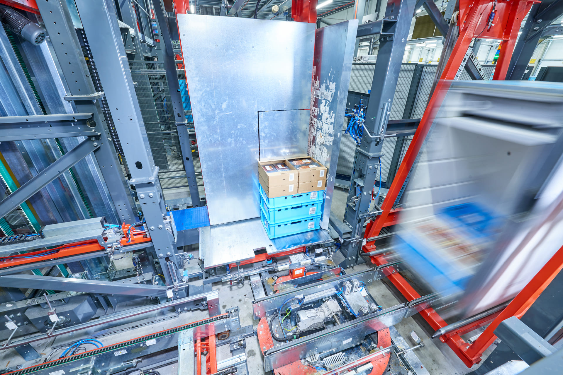 Ein Stapel blauer Kisten und Kartons liegt auf einer Plattform in einem automatisierten Lager, umgeben von Metallmaschinen und roten Rahmen - Maximale Frische durch maßgeschneiderte Automatisierung sorgt für effiziente Abläufe. Bewegungsunschärfe zeigt sich bei sich schnell bewegenden Geräten.