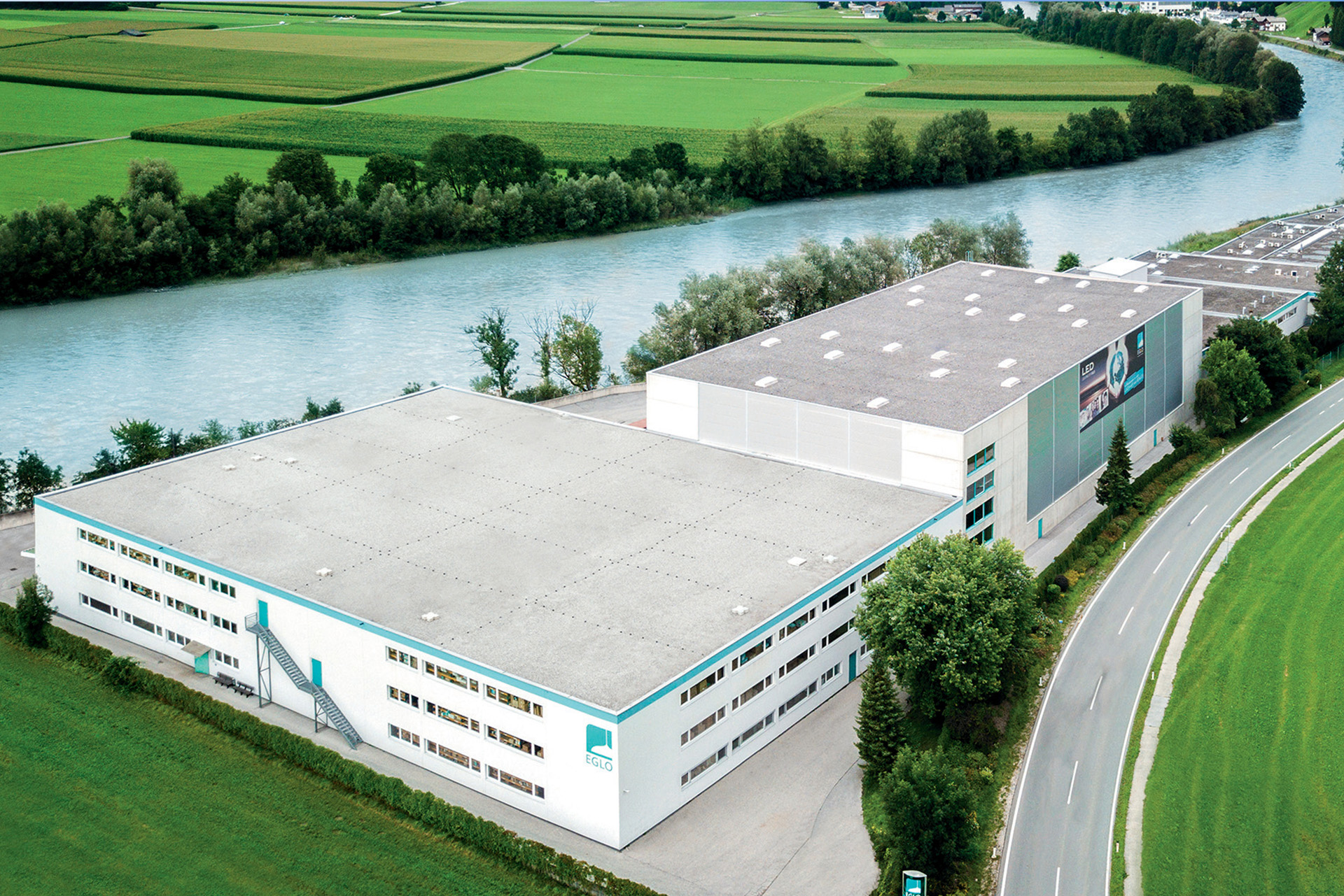 Un gran edificio industrial blanco con tejado plano está situado junto a un río y rodeado de campos verdes y árboles.